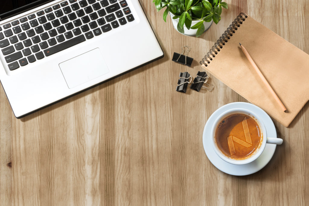 An image of a laptop, coffee, notebook, and plant on a wooden desk.  